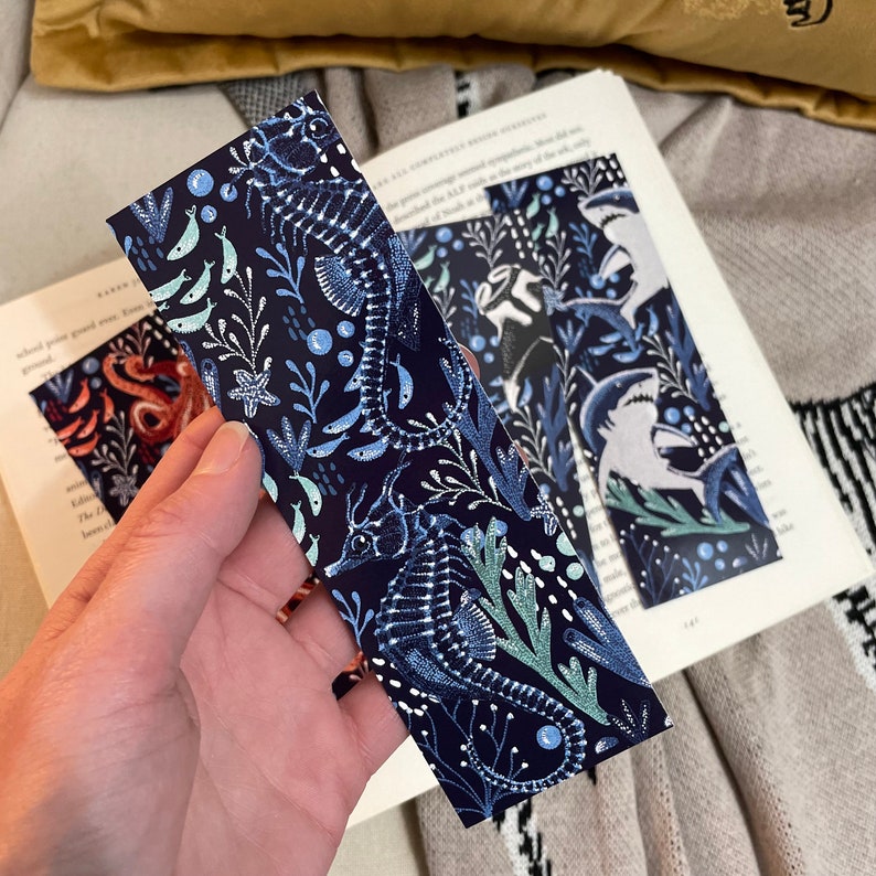 May include: A blue bookmark with a white and light blue seahorse design. The bookmark features a repeating pattern of seahorses, seaweed, and bubbles.