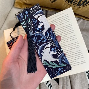 May include: A blue bookmark with a white tassel. The bookmark features a repeating pattern of white sharks and blue seaweed on a dark blue background.