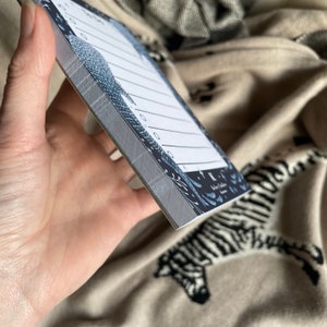 Puede incluir: Un peque&ntilde;o bloc de notas con un dise&ntilde;o estampado en azul y blanco. El bloc de notas tiene un lomo gris y p&aacute;ginas rayadas. El texto "Lost & Found" est&aacute; impreso en la parte delantera del bloc de notas.