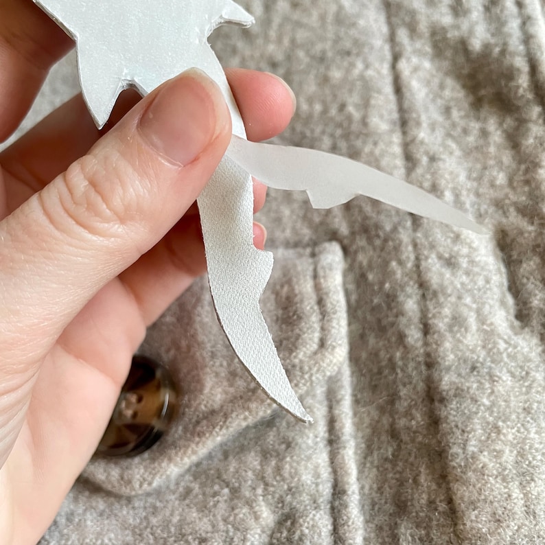 May include: A hand holding a white paper bird cutout, peeling it away from a light grey fabric. The fabric has a textured surface, and a brown button is visible in the background.