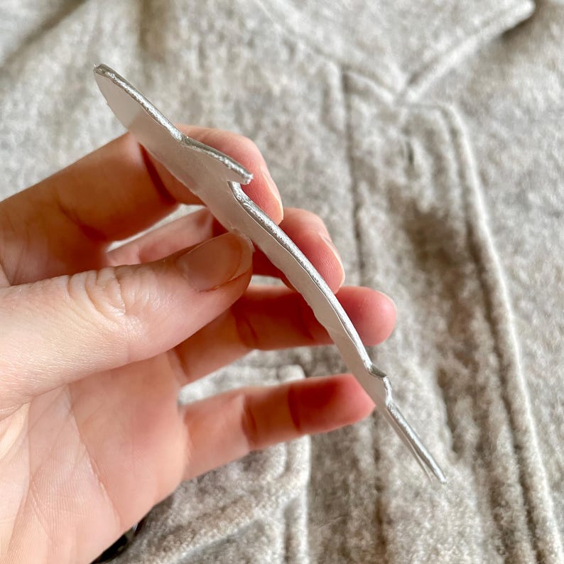 May include: A silver metal feather-shaped object. The feather has a textured edge and a stem. The object is held against a grey background.