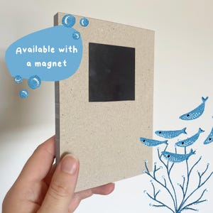 Puede incluir: Una tarjeta marr&oacute;n con un im&aacute;n cuadrado negro en la parte delantera. La tarjeta tiene una ilustraci&oacute;n azul y blanca de peces y algas en la parte posterior. El texto "Available with a magnet" est&aacute; en la parte delantera de la tarjeta.