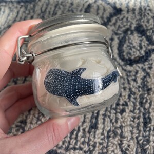 Puede incluir: Un tarro de vidrio con tapa blanca y cierre plateado. El tarro contiene una sustancia blanca y una pegatina de tibur&oacute;n ballena en blanco y negro.
