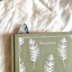 Peut inclure: Un livre vert intitul&eacute; "Persuasion" avec des illustrations de plumes blanches et un signet d&eacute;coratif. Le signet est de petite forme triangulaire avec un motif d&eacute;taill&eacute; dans les tons gris et blanc.