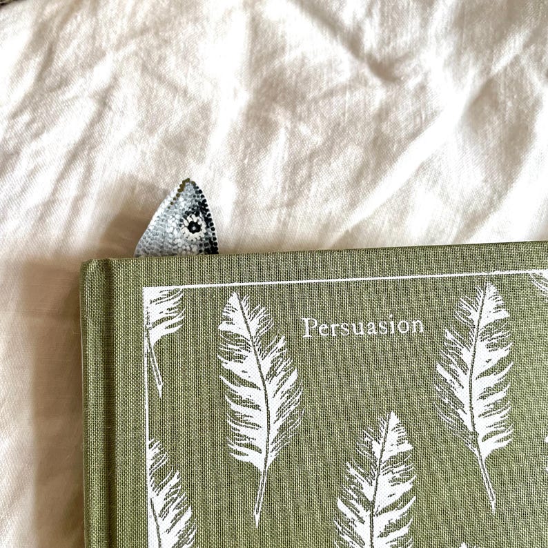 Peut inclure: Un livre vert intitul&eacute; "Persuasion" avec des illustrations de plumes blanches et un signet d&eacute;coratif. Le signet est de petite forme triangulaire avec un motif d&eacute;taill&eacute; dans les tons gris et blanc.