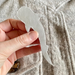 May include: A white shark-shaped cutout is held against a grey fabric background. The cutout is made of a light-coloured material and has a smooth texture. The shark's silhouette is clearly defined, with fins and a tail.