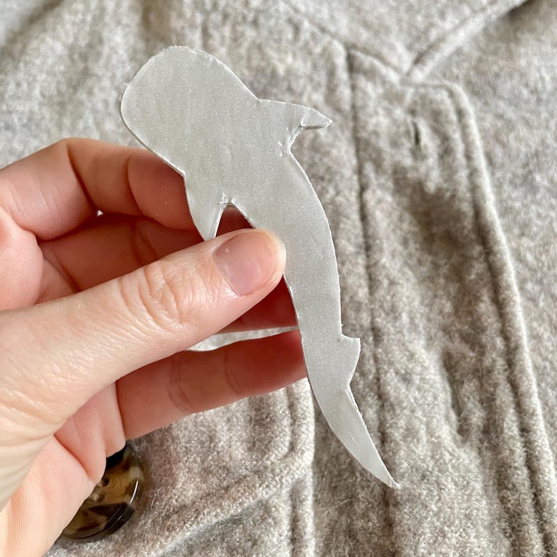 May include: A white shark-shaped cutout is held against a grey fabric background. The cutout is made of a light-coloured material and has a smooth texture. The shark's silhouette is clearly defined, with fins and a tail.