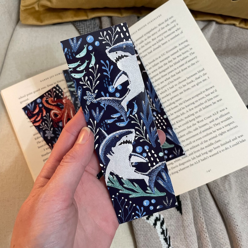 May include: A blue bookmark with a white background featuring a repeating pattern of white sharks, blue fish, and green seaweed. The bookmark is held in a hand.