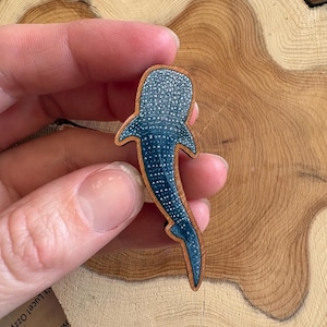 May include: A wooden whale shark brooch featuring a dark blue body with a pattern of white spots. The shark is depicted from a top-down perspective with a natural wood border outlining the shape of the creature, showcasing a smooth finish.
