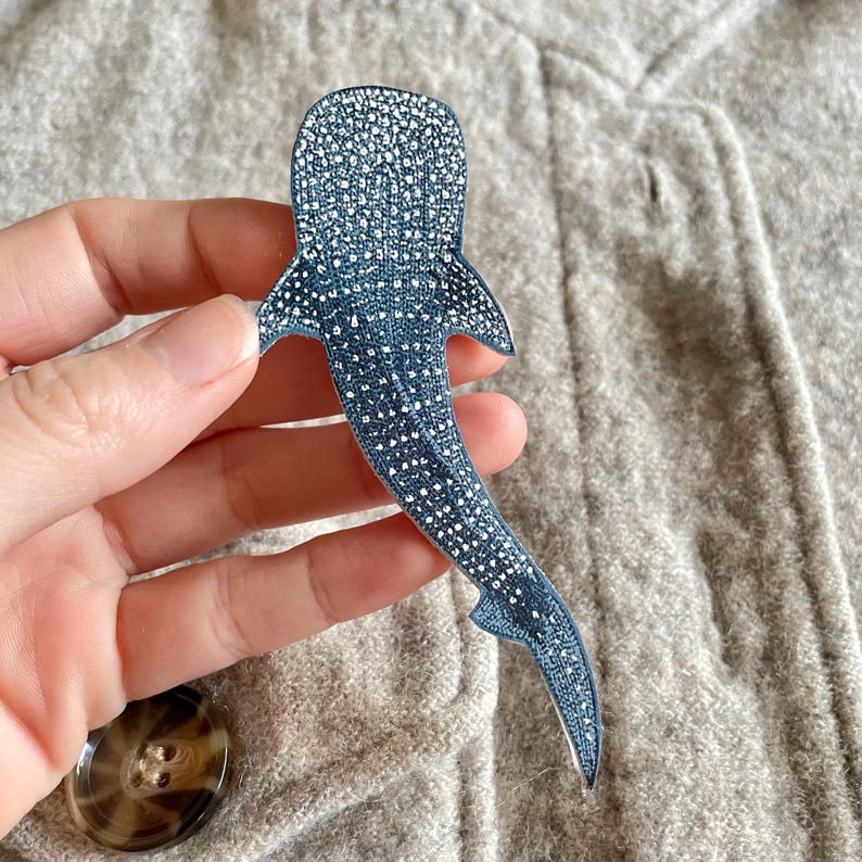 May include: A blue and white whale shark-shaped pin. The pin features a detailed pattern of white spots on a blue background. The pin is held against a grey fabric background.