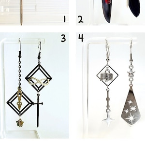 Könnte beinhalten: Vier Paar Hängeohrringe mit verschiedenen Anhängern, darunter ein Schwert, ein Unendlichkeitszeichen, ein Buch, eine Feder und Sterne. Die Ohrringe sind silber und schwarz.