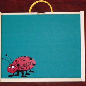May include: A turquoise magnetic dry erase board with a white frame and a yellow rope handle. The board has a red and black ladybug sticker with the name "Megan" written on it.