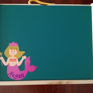 May include: A wooden framed chalkboard with a green surface. A pink mermaid with a crown and a wand is on the chalkboard. The mermaid's tail is purple and has a white wave pattern. The name "Megan" is written below the mermaid.