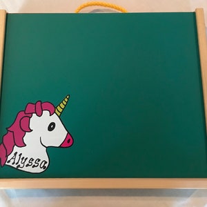May include: A green chalkboard with a light wood frame and a yellow rope handle. A cartoon unicorn with pink hair, a yellow horn, and the name "Alyssa" is on the lower left corner. The chalkboard is rectangular.