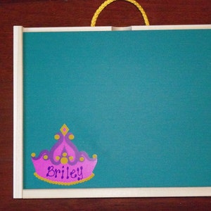 May include: A teal-colored chalkboard with a wooden frame and a yellow rope handle. A pink and purple crown with the name "Briley" is affixed to the lower right corner. The chalkboard is designed for writing or drawing.