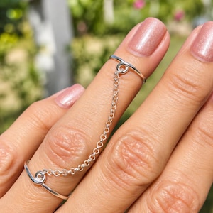 May include: A silver chain ring set consisting of two thin, smooth bands connected by a delicate, fine-link silver chain. Each band features a small, circular loop where the chain attaches, creating a continuous link between the two rings.