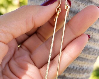May include: A pair of long, slender gold-tone drop earrings featuring a minimalist bar design. One earring has a textured, hammered finish that catches the light, while the other earring features a smooth, polished surface. Both hang from gold-tone hooks.