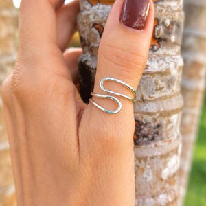 May include: A silver-colored ring featuring a continuous, open-ended design that forms a stylized S-shape. The band is crafted from a smooth, polished metal wire that wraps around the finger, creating two parallel loops that remain open.