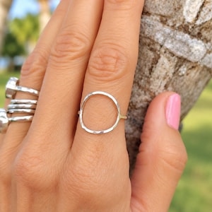 May include: A silver ring featuring a thin, hammered, organic circular band on the middle finger, accompanied by three additional silver rings with textured, irregular surfaces stacked on the ring finger, all displaying a polished metallic finish.