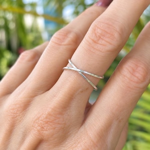 May include: A silver-colored ring featuring a thin, polished band that crosses over itself in an X-shape design. The metal has a smooth, reflective surface that catches the light, creating a simple and clean aesthetic on the finger.