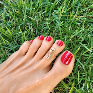 May include: A gold-toned metal toe ring featuring a double spiral design. The ring is worn on the second toe, which is positioned next to a large toe with bright red nail polish. The metal band is open-ended, showcasing a simple, polished finish.