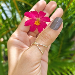 May include: A polished silver ring featuring a simple crossover design with two thin bands that intersect at the center. The ring is worn on a thumb, positioned below a vibrant magenta flower with a yellow center, set against a blurred green background.