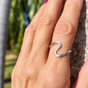 May include: A polished silver ring featuring a single continuous wave-shaped band that curves upward and downward. The metal has a smooth, reflective surface that catches the light, creating a simple and clean aesthetic for this finger accessory.