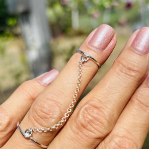 May include: A silver finger chain ring consisting of two thin, smooth bands connected by a delicate, small-linked silver chain. The bands feature a simple, open-ended design where the metal curves slightly at the connection point for the chain.