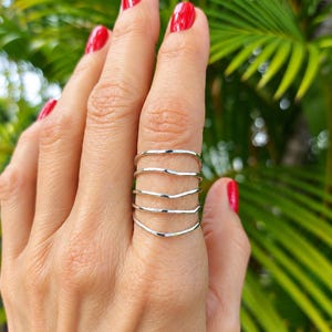 May include: A silver ring featuring five thin, wavy bands that are connected to form a single piece. The metal has a polished, reflective finish, and the design consists of multiple horizontal, undulating lines that wrap around the finger.