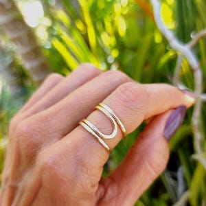 May include: A gold-colored ring with a unique U-shaped design. The ring is worn on a finger, with a blurred green background. The ring appears to be made of metal and has a smooth, polished finish. Jewelry.