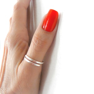 May include: A simple silver ring with a bypass design, featuring two parallel bands that overlap slightly. The ring is crafted from a smooth, polished metal with a reflective finish, creating a clean and understated look on the finger.