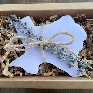 May include: A purple Texas-shaped candle with a bundle of lavender tied with twine. The candle is sitting in a box filled with brown packing paper.