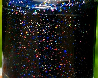 May include: A bottle of nail polish with a dark, glitter-filled formula. The polish appears black with a multitude of colourful, iridescent glitter particles. The bottle has a white cap. The background is a blurred green, suggesting an outdoor setting.