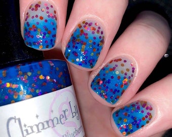 May include: Close-up of a hand with manicured nails. The nails feature a blue glitter gradient design with a mix of colorful glitter. A bottle of "Glimmer by Blueberry" nail polish is visible in the image.