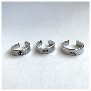 Puede incluir: Tres anillos de plata con diferentes diseños. El primer anillo tiene un diseño de árbol, el segundo anillo tiene un diseño de arcoíris y el tercer anillo tiene un diseño de montaña.