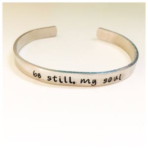 Puede incluir: Un brazalete de plata con las palabras "be still, my soul" grabadas.