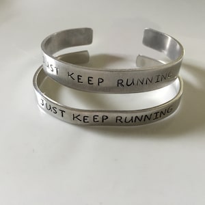 Pode incluir: Duas pulseiras de prata com as palavras "Just Keep Running" gravadas.