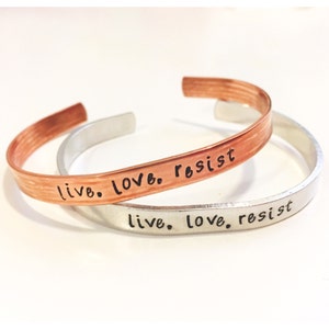 Puede incluir: Dos pulseras de metal con las palabras "live. love. resist" grabadas en ellas. La pulsera superior es de cobre y la inferior es de plata.