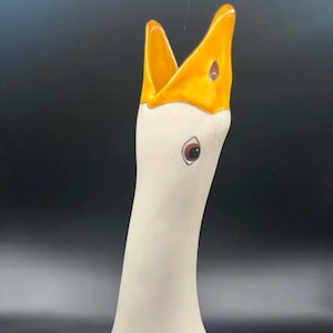 May include: A ceramic goose figurine with a long neck and an open beak. The body is off-white with light blue stippled feather patterns. The beak is bright yellow, and the eye is dark brown with a black pupil, set against a smooth, glossy finish.