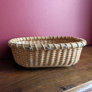 May include: An oval-shaped woven basket made of light-colored natural reed or wood splints. The basket features a reinforced rim with a decorative lashing pattern and small, evenly spaced metal fasteners securing the rim to the body structure.