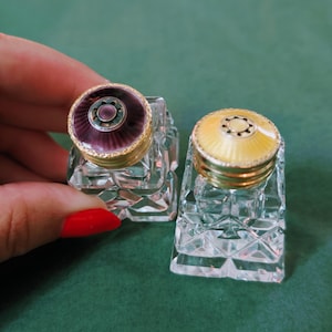 May include: Two antique glass inkwells with ornate, colorful lids. One lid is purple and gold, the other is yellow and gold. The inkwells are clear and faceted, reflecting light. A classic desk accessory.
