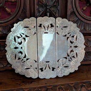 Könnte beinhalten: Ein silberfarbener Metalluntersetzer mit dreiteiligem Klappdesign und filigranen, ausgeschnittenen Blumen- und Muschelmotiven. Die Oberfläche hat ein poliertes, reflektierendes Finish mit gewellten Rändern und symmetrischen Mustern.