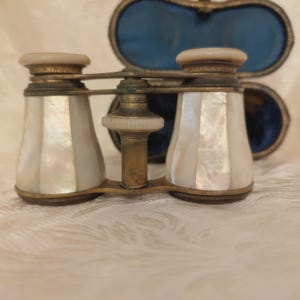 May include: Antique opera glasses with a mother-of-pearl finish and brass accents. The binoculars are in a gold-colored frame, with a cream-colored top and a pearlescent white body. A blue velvet case is in the background.