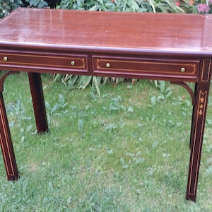 May include: A mahogany desk with two drawers and gold accents. The desk has a dark wood finish and intricate inlays. It is sitting on a green lawn.
