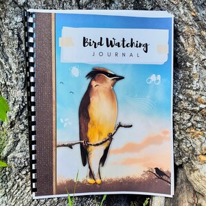 Puede incluir: Un diario de espiral con una portada que muestra un ampelis americano posado en una rama contra un cielo azul y naranja. El texto "Bird Watching JOURNAL" está impreso en la parte superior. La portada incluye ilustraciones de un escarabajo y binoculares.