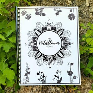 Può includere: Un diario con rilegatura a spirale bianca e un design a inchiostro nero che presenta un motivo floreale centrale in stile mandala e il testo My Wildflower Journal. La copertina include varie illustrazioni a silhouette nera di fiori selvatici.