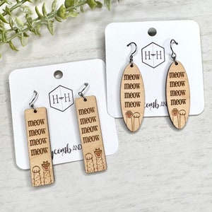 Op de afbeelding: Twee paar houten oorbellen met een kattenpootontwerp en het woord "meow" dat meerdere keren wordt herhaald. De oorbellen zijn op een witte achtergrond.