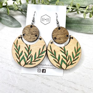 May include: Wooden earrings featuring a two-part design. The top is a small, round, dark brown piece, and the bottom is a light brown, crescent-shaped piece with green leaf details. The earrings hang from silver-coloured hooks.