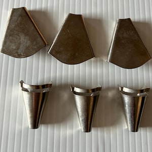 May include: A collection of six silver-toned metal components arranged in two rows. The top row features three flat, trapezoidal-shaped pieces, while the bottom row contains three conical, funnel-shaped pieces with open tops and small side tabs.