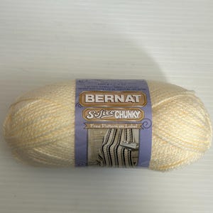 May include: A skein of Bernat Softee Chunky yarn in a creamy off-white color. The label features the brand name and the text "Softee Chunky" with a graphic of a knitted item. The label also states "Free Pattern on Label".